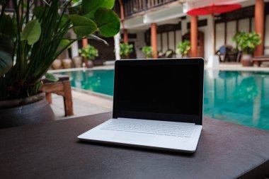 Uzaktan kumandalı dijital bir göçebenin dizüstü bilgisayarı oteldeki güzel bir havuzun arka planına karşı. Özgürlük, uzaktan çalışma, serbest çalışma, teknoloji, internet, seyahat ve tatil kavramları