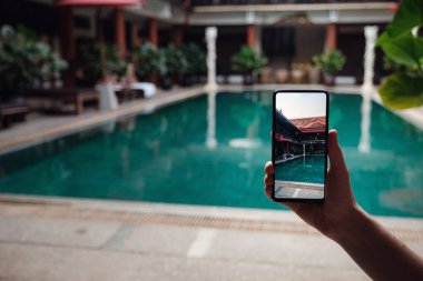 Oteldeki havuzun arka planında el ele tutuşan telefon. Ekrandaki fotoğraf makinesi. Kapat elini akıllı telefon yaz tatilinin fotoğrafını çek..