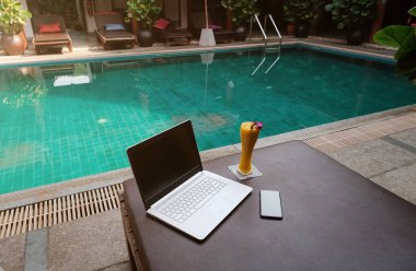 Beyaz bir dizüstü bilgisayar, akıllı telefon ve yüzme havuzuna karşı güneşli bir yatakta mango smoothie. Yeni bir günün başlangıcı. Serbest iş konsepti. Esnek uzaktan çalışma, seyahat, reklam ve kopyalama alanı