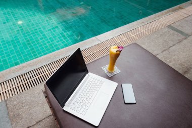 Beyaz bir dizüstü bilgisayar, akıllı telefon ve yüzme havuzuna karşı güneşli bir yatakta mango smoothie. Yeni bir günün başlangıcı. Serbest iş konsepti. Esnek uzaktan çalışma, seyahat, reklam ve kopyalama alanı