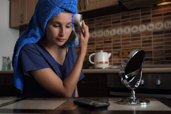 A woman is sitting in the kitchen and doing a saved procedure using a beauty gadget. Hardware cosmetology and personal care, Skin care and age cosmetics concept