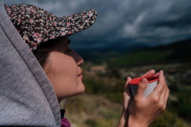 Şapkalı turist kız elinde sıcak bir içecekle bardağı tutuyor ve manzaranın tadını çıkarıyor. Güzel yeşil vadi ve dramatik yağmur gökyüzü