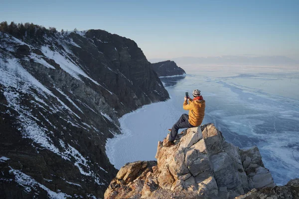 Lago Baikal in inverno. Uomo seduto su una scogliera e scattare foto su smartphone . — Foto Stock