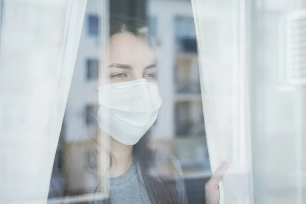 脸上戴面具的年轻女人从窗户往外看 在检疫室待在家里 — 图库照片