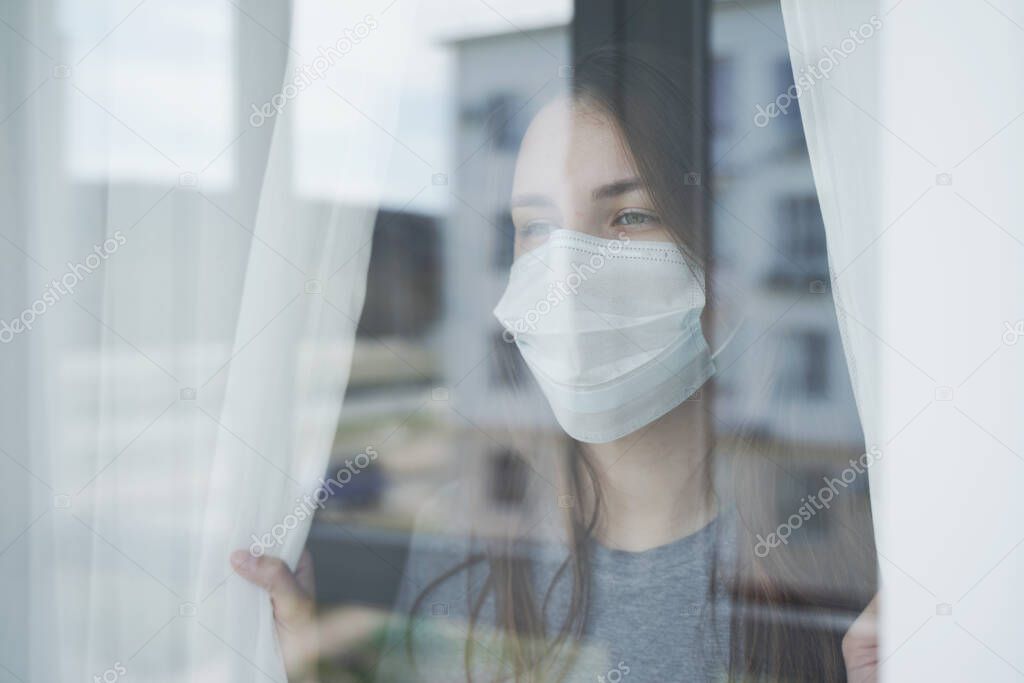 Young Woman in Face Mask Looking out the Window. Staying Home in Self-Quarantine.
