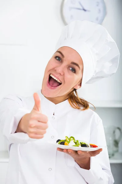 Girl in chef's hat and white coat showing salad — Stock Photo, Image