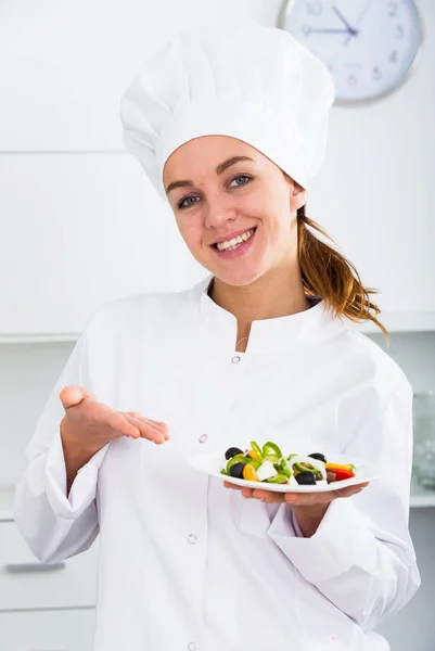 Girl in chef's hat and white coat showing salad — Stock Photo, Image