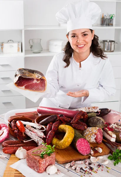 Dziewczyna gotować kosztuje w pobliżu stołu, na którym kiełbasy i wędzone mięso — Zdjęcie stockowe