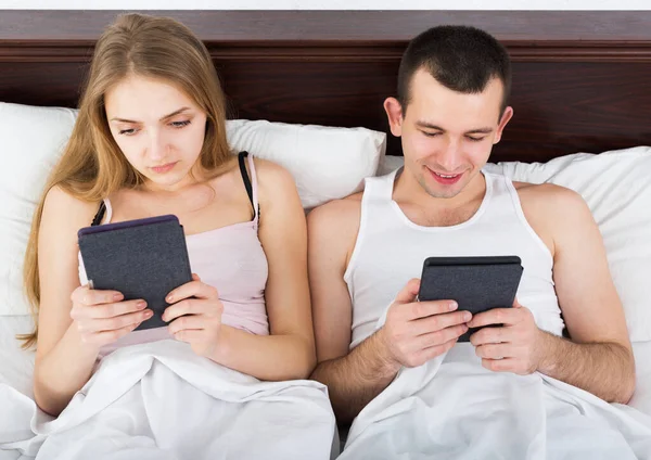 Молодая пара читает новости из электронных читателей в постели — стоковое фото