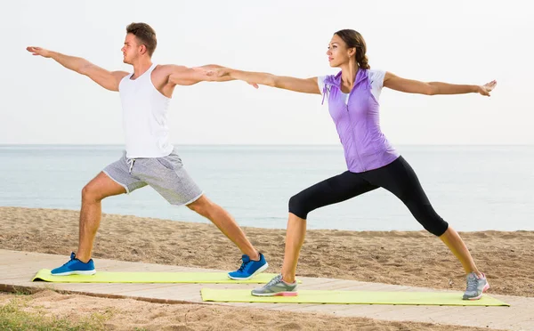 Young couple exercising yoga poses standing on sunny beach by ocean in morning