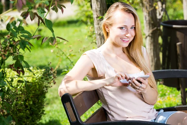 明るいですブロンド若いです女性テキストを使用して携帯電話で公園で晴れた日 — ストック写真
