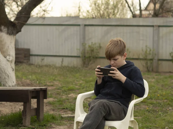 Vit Blond Pojke Spelar Smartphone Medan Sitter Vit Plaststol Trädgården — Stockfoto