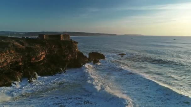 ナザレ ポルトガル 4Kの日没時に大きな波と大西洋の海岸に要塞と崖の上の灯台の周りの空中 — ストック動画