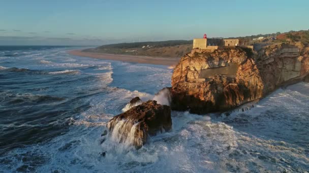 ナザレ ポルトガル 4Kの日没時に大きな波と大西洋の海岸に要塞と崖の上の灯台の航空写真 — ストック動画