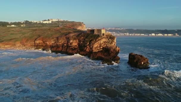 ナザレ ポルトガル 4Kの日没時に大きな波と大西洋の海岸に要塞と崖の上の灯台の周りの空中 — ストック動画