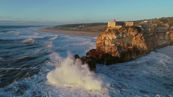 ポルトガルのナザレの日没時に大きな波を持つ大西洋の海岸沿いの要塞と崖の上の灯台の空中ビュー スローモーション4K — ストック動画