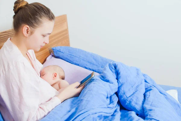 Mulher usando telefone celular na cama durante amamenta bebê recém-nascido — Fotografia de Stock