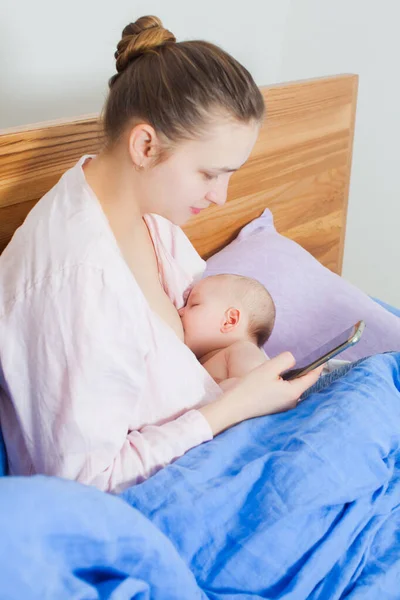 Permiso de maternidad. Madre viendo video en smartphone y amamantando a su pequeño bebé — Foto de Stock