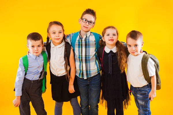 Chytré školáci se usmívají a dívají se na kameru přes žluté pozadí. Šťastné děti ve školní uniformě s batohy — Stock fotografie