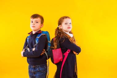 Schoolgirl and schoolboy with backpacks and crossed hands standing over yellow background