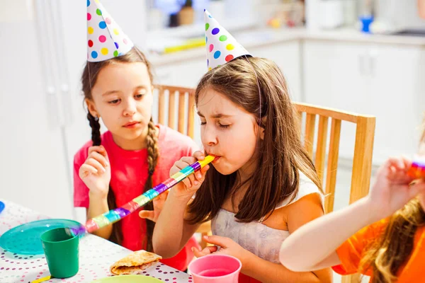 Klein mooi meisje probeert een fluitje van een cent te blazen op het feest. Zusters zitten aan de verjaardagstafel — Stockfoto