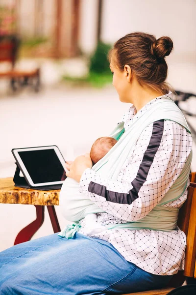 Young mother with little baby in sling sitting at cafe outdoors and working using tablet — 스톡 사진