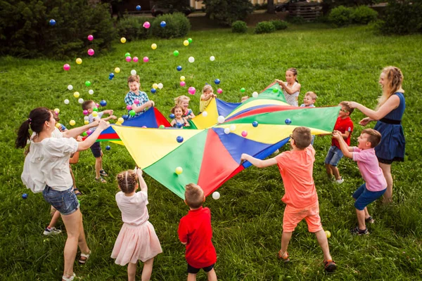 Счастливые дети, размахивающие радужным парашютом, полным мячей — стоковое фото