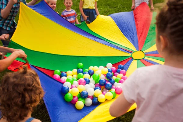 Niños felices agitando paracaídas de arco iris lleno de bolas —  Fotos de Stock