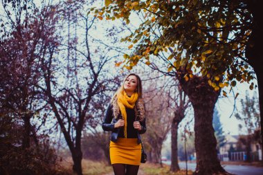Portrait of young woman wearing black leather jacket and yellow scarf outdoors. Pretty girl walking in autumn park.