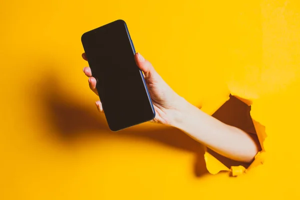 Mano femenina fuera del agujero en la pared de papel, sosteniendo el teléfono inteligente con una pantalla negra . — Foto de Stock
