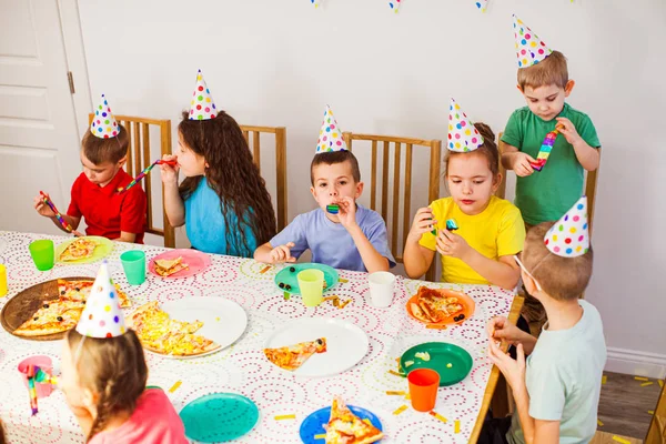 Kedves gyerekek, akik szülinapi sapkát viselnek, és jól érzik magukat együtt. A gyerekek finom pizzát esznek a partin. — Stock Fotó