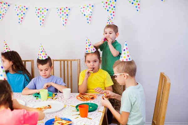 Radostné děti jedí pizzu u velkého stolu. Děti nosí narozeninové klobouky, baví se spolu. — Stock fotografie