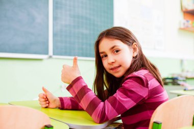 Primary schoolgirl with thumb up in empty classroom