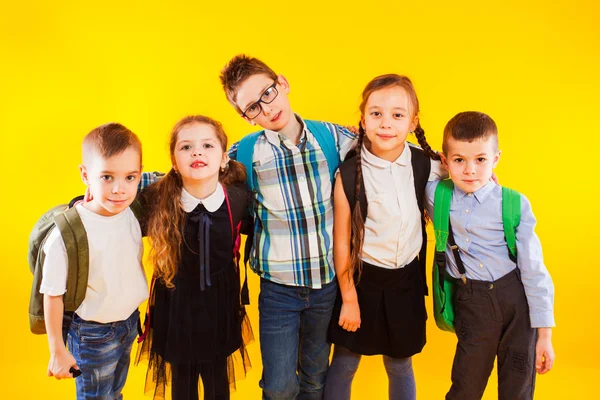 Egy csapat boldog gyerek egyenruhában, iskolatáskákkal ölelkezve és kamerába nézve sárga háttér felett — Stock Fotó