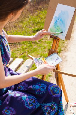 Girl is painting with watercolor on the easel outdoors. Spring imagination.
