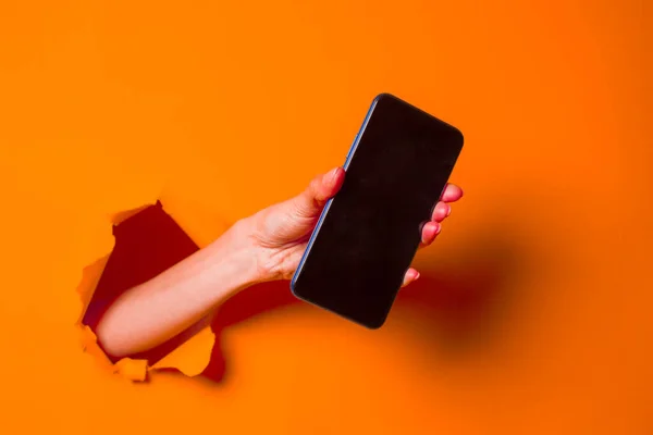 Una mujer sostiene un teléfono a través de un fondo de papel naranja andrajoso, primer plano . — Foto de Stock