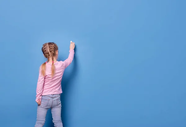 Kleines Mädchen schreibt mit Kreide auf blauem Hintergrund — Stockfoto