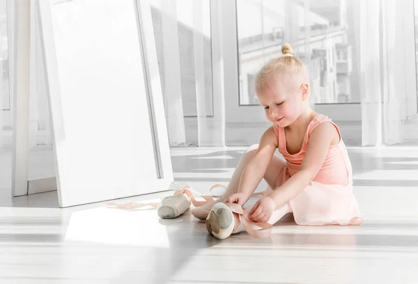 Cute little blonde girl sitting on the floor and tying ballet shoes