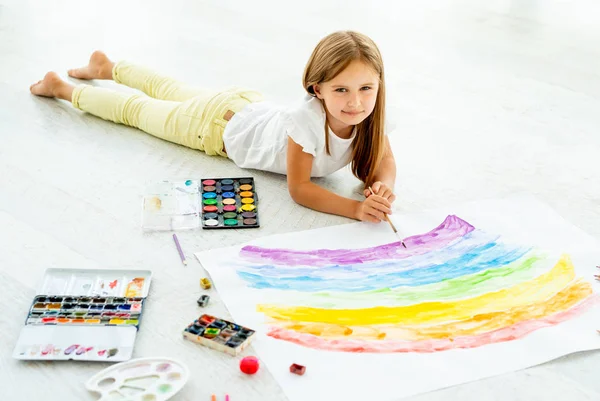 Маленька дівчинка малює лежачи на підлозі Маленька весела дівчинка малює лежачи на підлозі — стокове фото