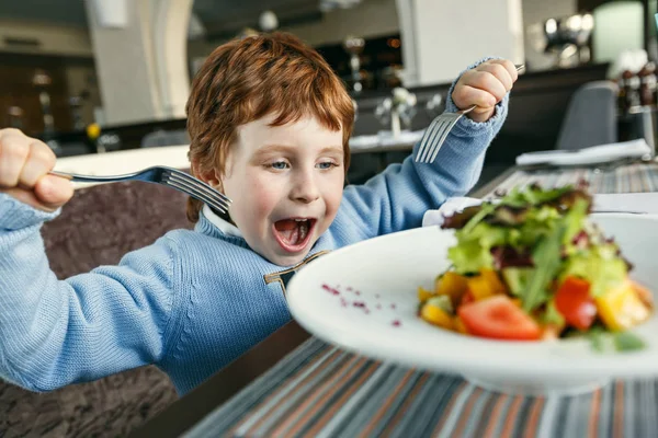 Niño pelirrojo con tenedores comiendo ensalada Imagen de archivo
