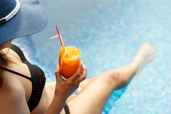 戴着蓝色夏帽 双腿细长的女人在游泳池边喝着鸡尾酒放松 — 图库照片