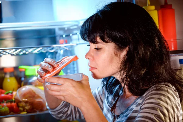 Mulher abriu a geladeira e cheira um recipiente de comida — Fotografia de Stock