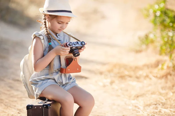 A little girl with an old camera on a country road sitting on a suitcase — Stock Photo, Image