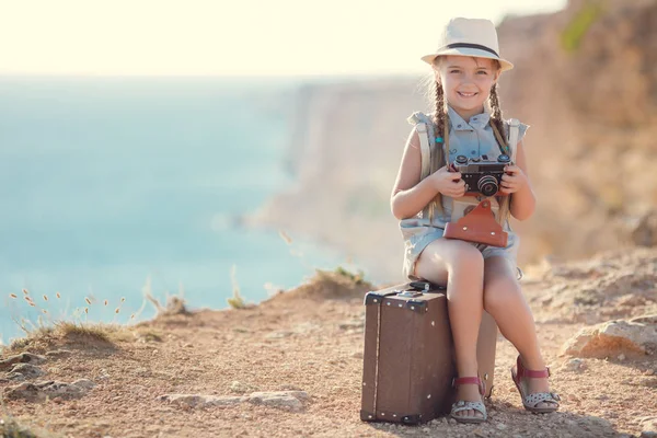 Una ragazzina con una vecchia macchina fotografica su una strada di campagna seduta su una valigia — Foto Stock