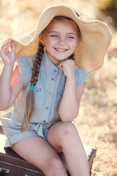 Klein meisje in een stro hoed, rust, zittend op een koffer — Stockfoto