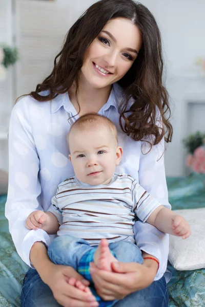 Young beautiful mother holding baby son on her lap, sitting on a bed — Stock Photo, Image
