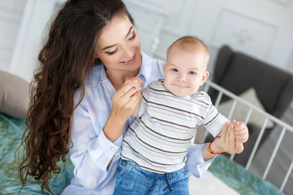 Young beautiful mother holding baby son on her lap, sitting on a bed — Stock Photo, Image