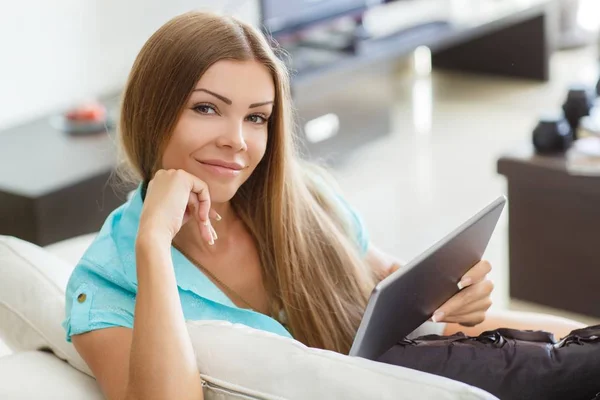 Hermosa mujer joven en ropa casual está utilizando una tableta digital y sonriendo mientras está sentado en el sofá en casa —  Fotos de Stock