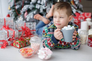 5 yaşında bir çocuk büyük bir fincan kahve marshmallow ile tutan güzel bir Noel ağacının yanındaki yatıyor