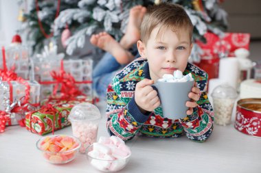 5 yaşında bir çocuk büyük bir fincan kahve marshmallow ile tutan güzel bir Noel ağacının yanındaki yatıyor